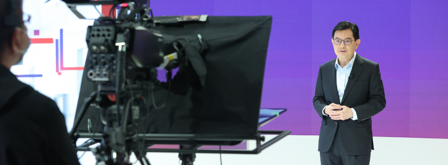 Man in suit speaks, camera recording; purple backdrop. Crew member visible in foreground.